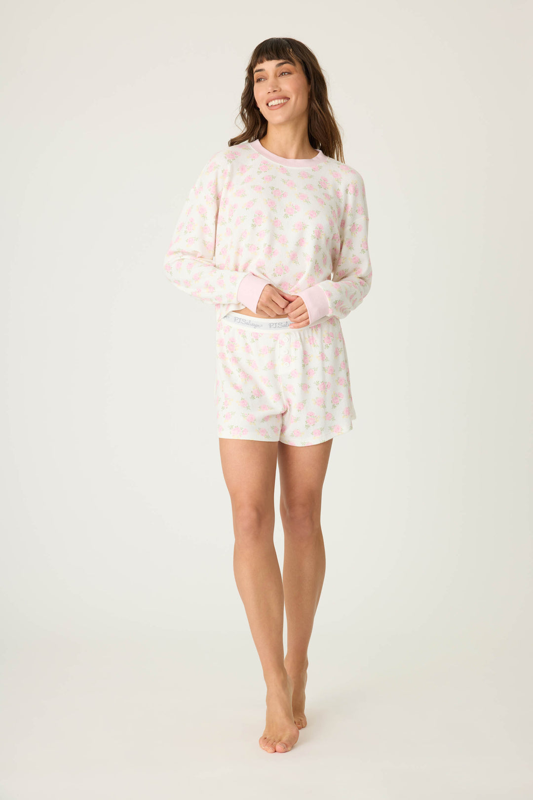 A woman stands barefoot against a plain background, smiling and looking slightly to the side, wearing the P.J. Salvage Boyfriend Boxer Ditsy Floral Shorts Set—a light pink, long-sleeve pajama top and matching floral print shorts.