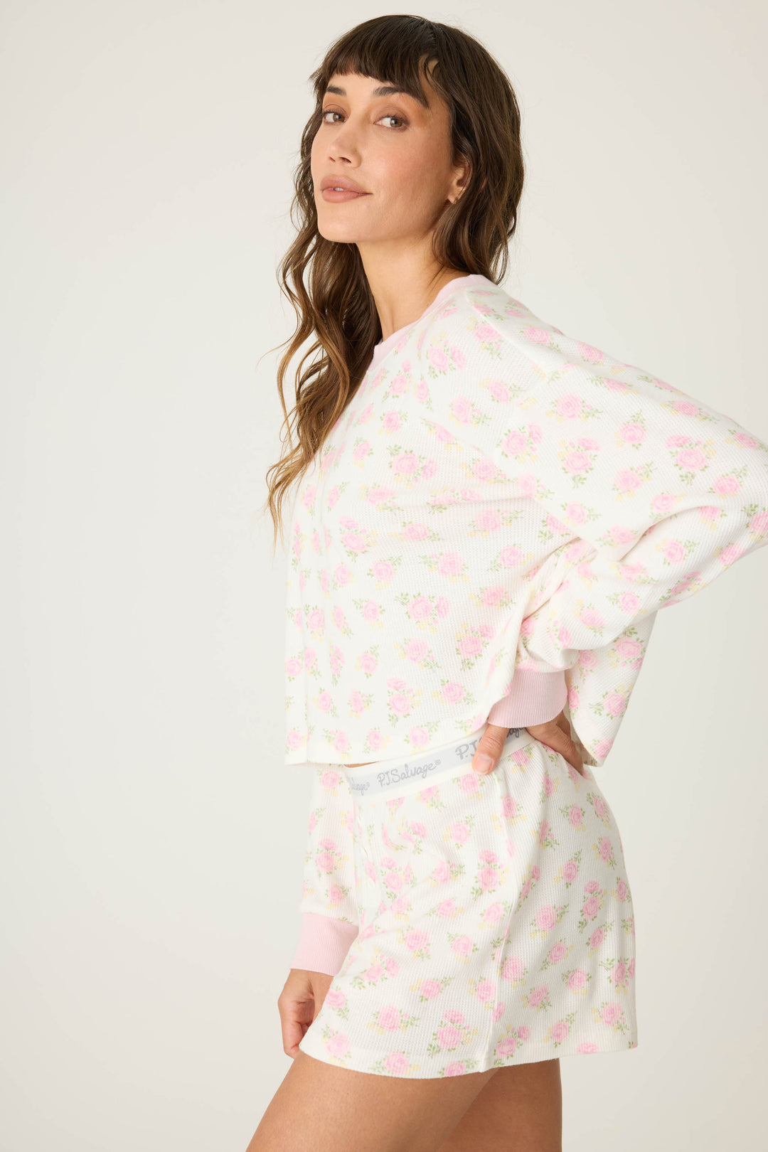 A woman stands sideways, looking at the camera, wearing the P.J. Salvage Boyfriend Boxer Ditsy Floral Shorts Set—a matching long-sleeve top and shorts with a light pink floral pattern on white—against a plain, neutral background.