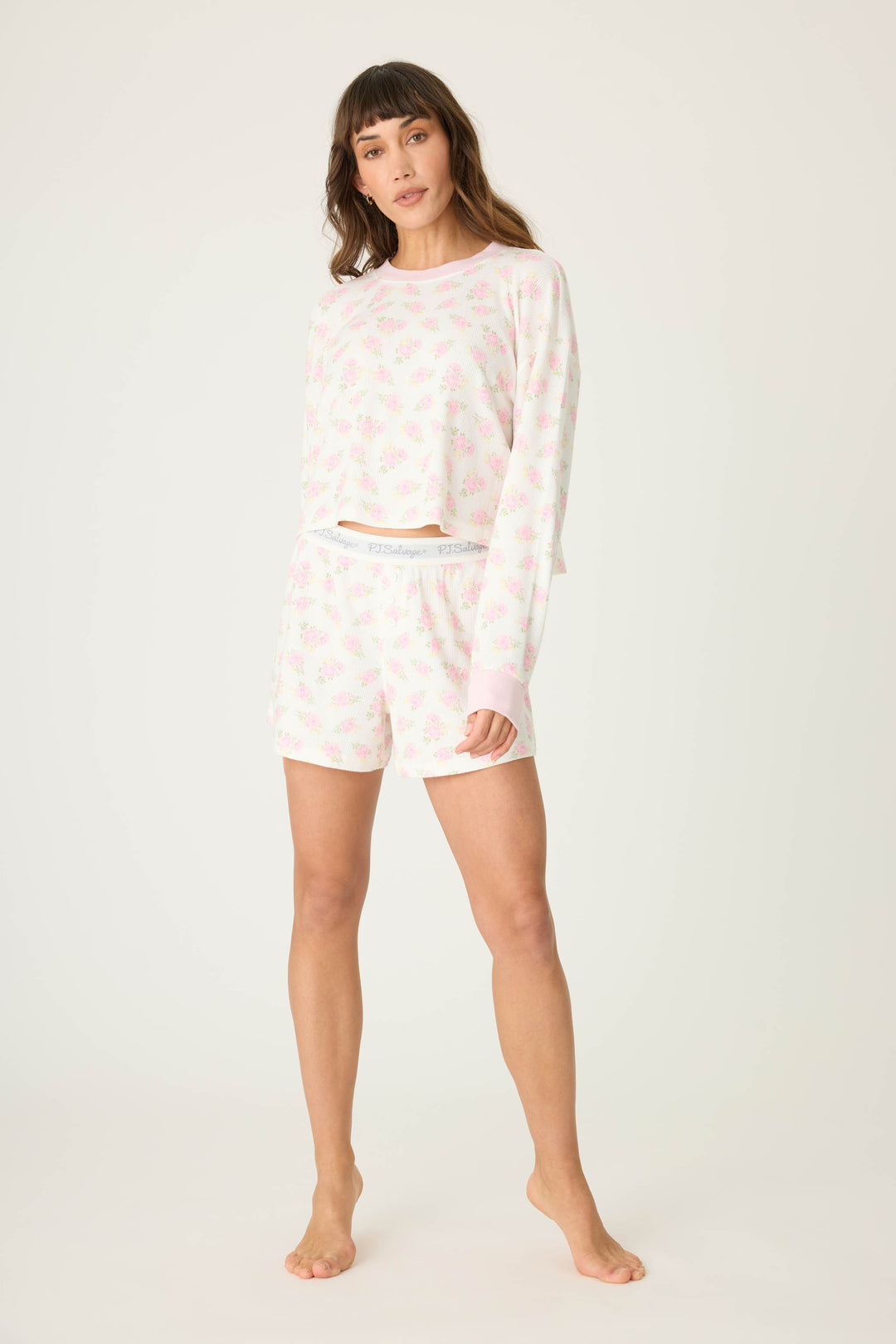 A woman stands barefoot against a plain background, wearing the P.J. Salvage Boyfriend Boxer Ditsy Floral Shorts Set, which features a matching long-sleeve top and shorts with a light pink floral pattern.
