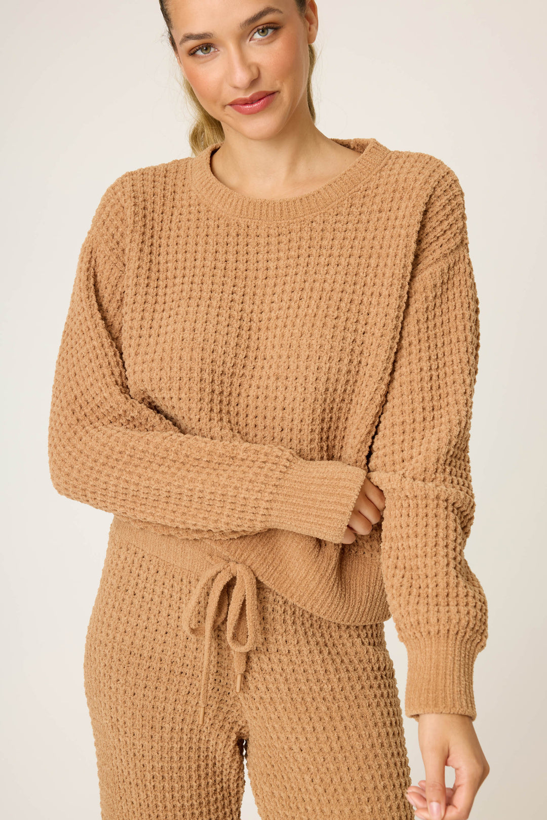 A woman in the P.J. Salvage Chenille Waffle Solid Caramel Sweater stands against a plain background, smiling slightly with her arms crossed.