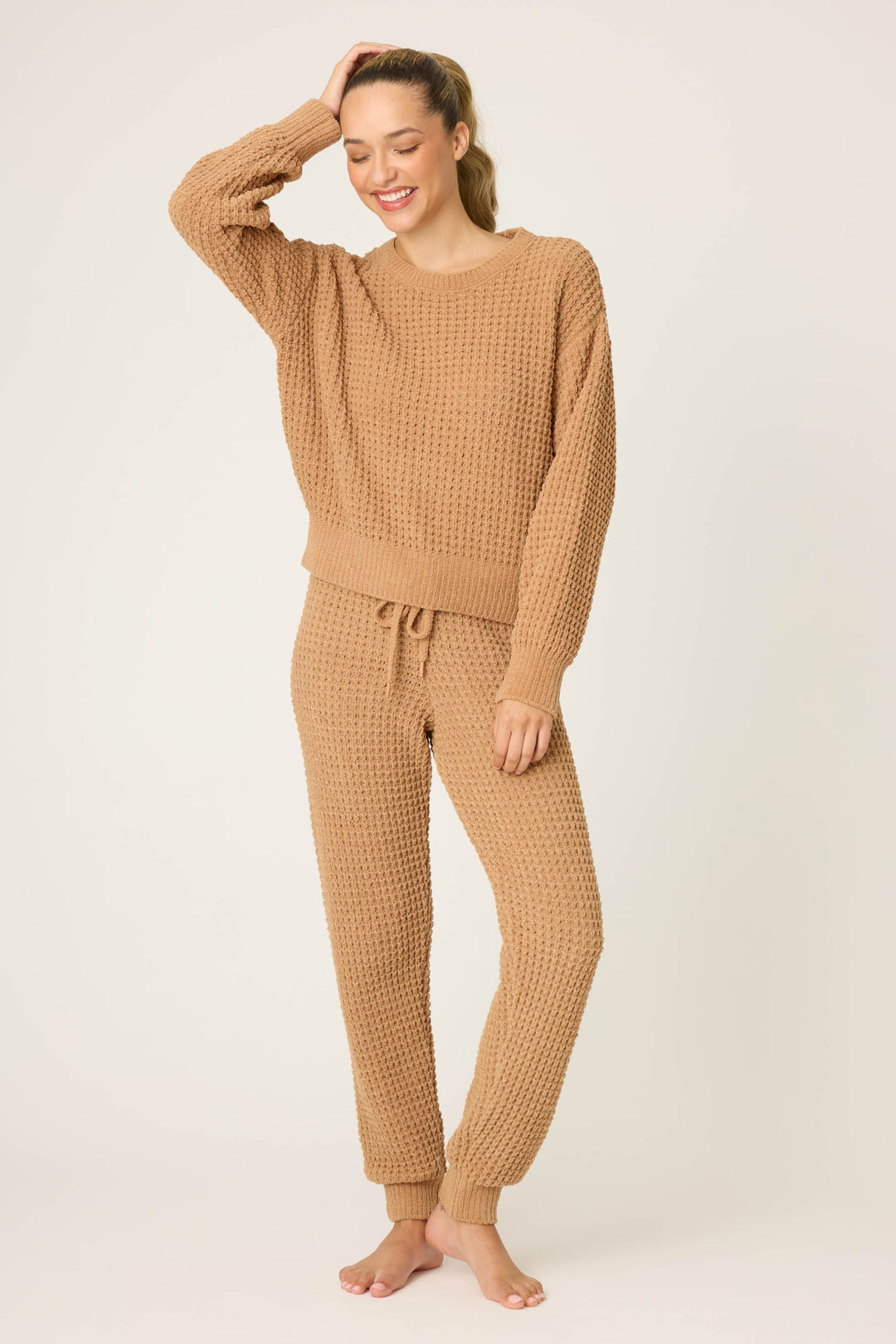 A person stands barefoot against a plain background, smiling and touching their head while wearing the P.J. Salvage Chenille Waffle Solid Caramel Sweater. Their other hand is relaxed by their side.