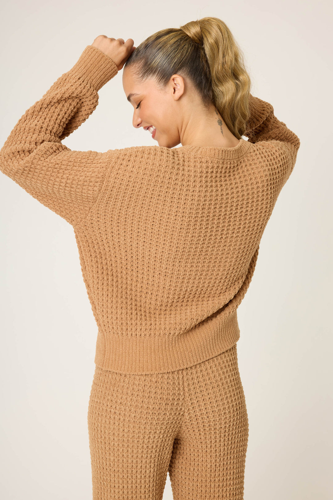 A person with a blonde ponytail wears the P.J. Salvage Chenille Waffle Solid Caramel Sweater and matching pants, facing away with raised arms and a slight smile against a plain, light-colored background.