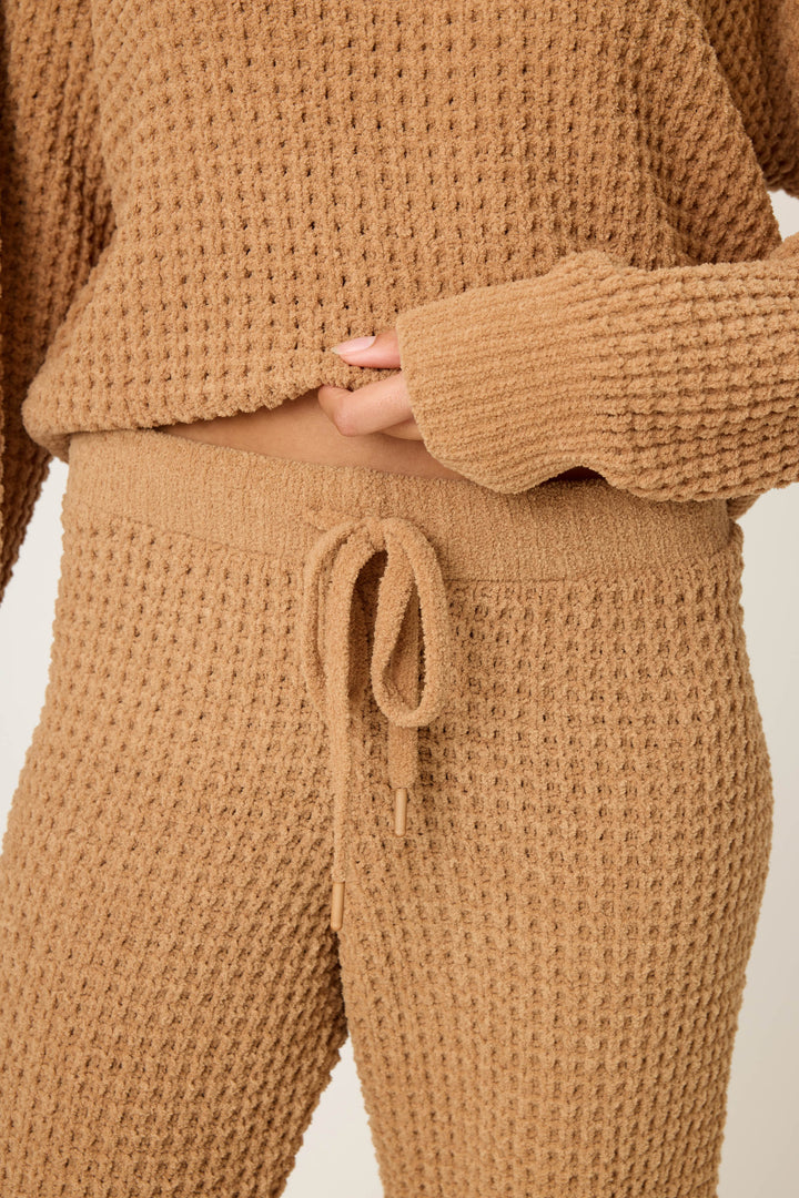 A person wears P.J. Salvage's Chenille Waffle Solid Caramel Banded Pant with a matching long-sleeve top, both in a textured waffle-knit fabric. Their hand is partly tucked under the top's hem.