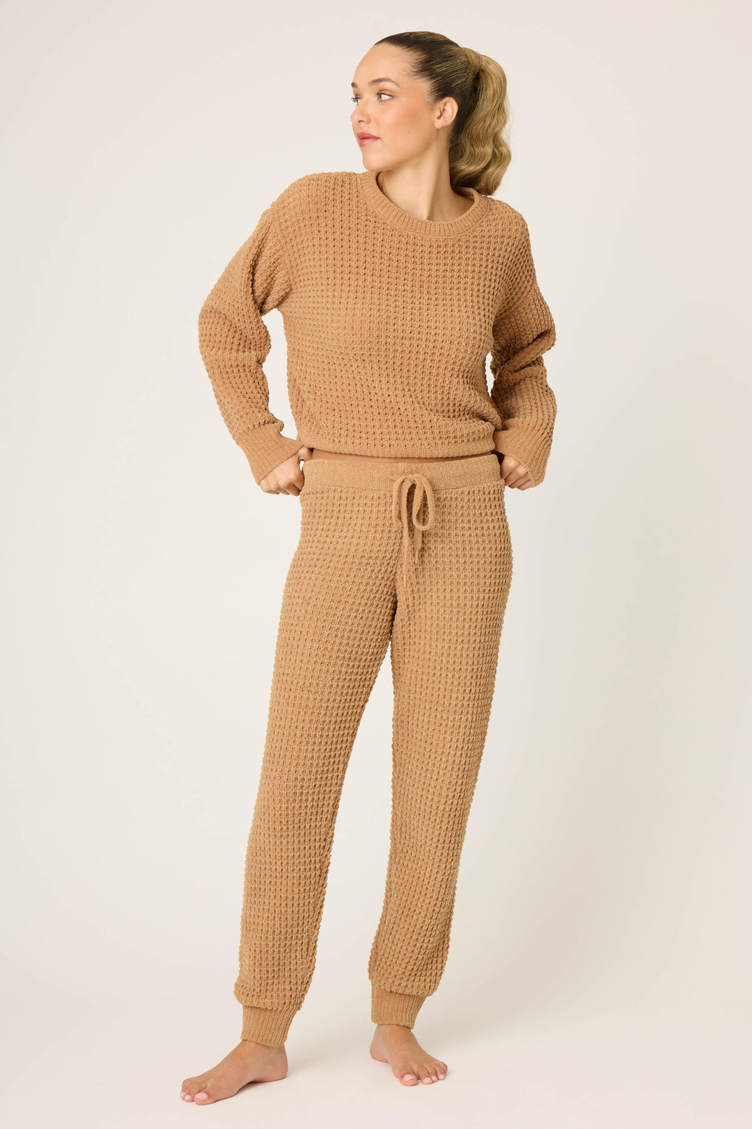 A woman stands barefoot against a plain background, wearing the P.J. Salvage Chenille Waffle Solid Caramel Banded Pant with a matching textured sweater set. She looks off to the side, hands on her hips.