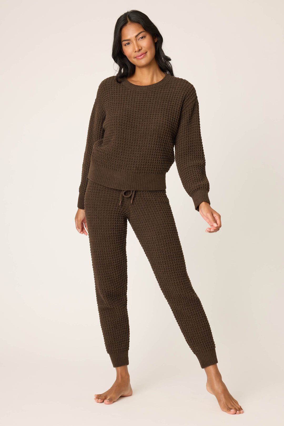 A person stands barefoot against a plain off-white background, wearing P.J. Salvage's Chenille Waffle Solid Chocolate Banded Pant with a matching long-sleeve textured sweater. They look relaxed, with one hand slightly raised.