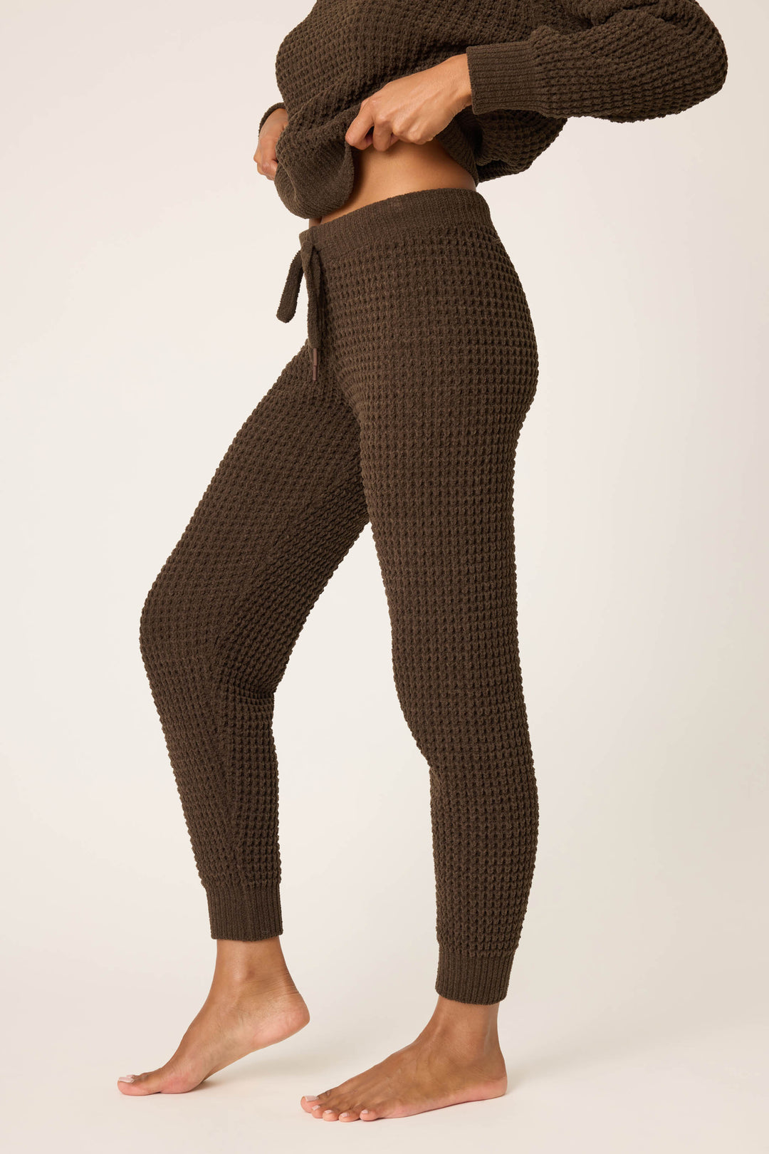 A person stands barefoot in P.J. Salvage’s Chenille Waffle Solid Chocolate Banded Pant, adjusting the waistband. They wear a matching long-sleeve top against a plain, light-colored background.