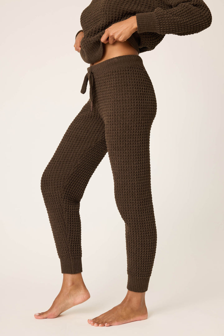 A person stands barefoot in P.J. Salvage’s Chenille Waffle Solid Chocolate Banded Pant, adjusting the waistband. They wear a matching long-sleeve top against a plain, light-colored background.