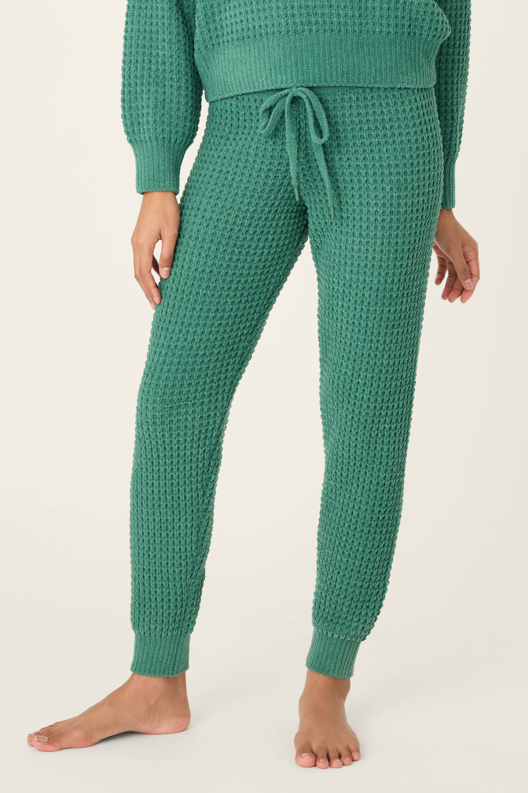 A person stands barefoot in P.J. Salvage's Chenille Waffle Solid Forest Banded Pant, featuring a drawstring waist and ribbed cuffs, paired with a matching knit sweater against a plain light background.