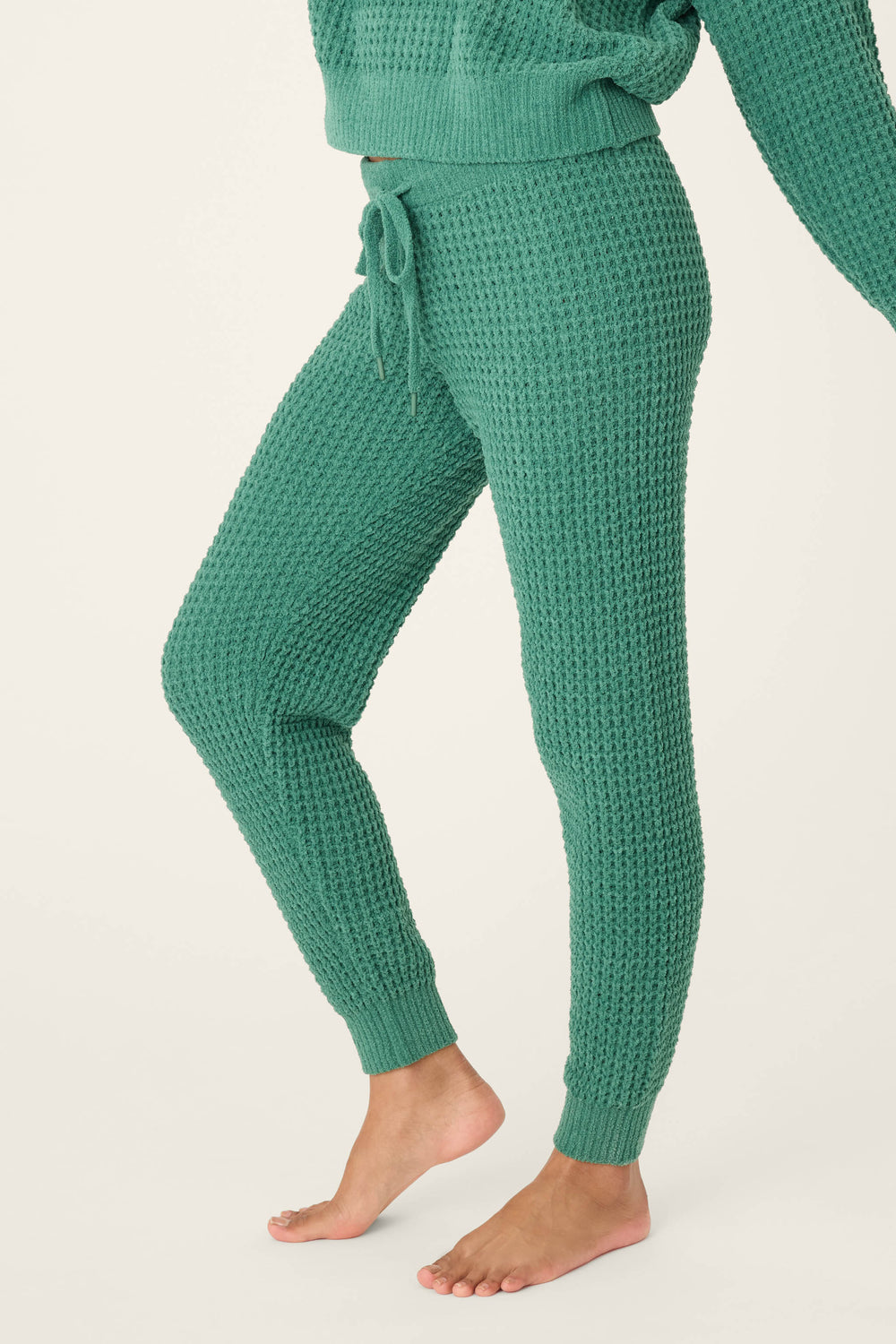 A person stands barefoot in P.J. Salvage Chenille Waffle Solid Forest Banded Pants, featuring textured green knit fabric and a drawstring waist. Part of a matching sweater is visible, with a plain off-white background behind them.