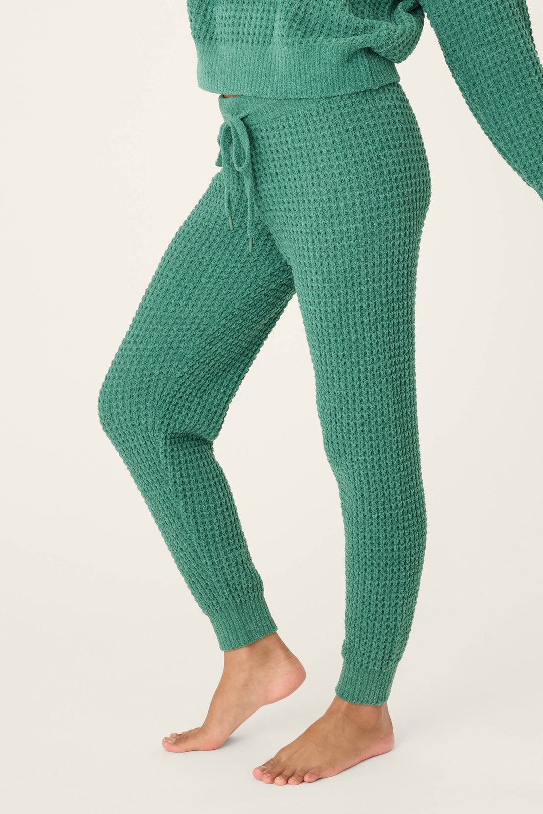 A person stands barefoot in P.J. Salvage Chenille Waffle Solid Forest Banded Pants, featuring textured green knit fabric and a drawstring waist. Part of a matching sweater is visible, with a plain off-white background behind them.