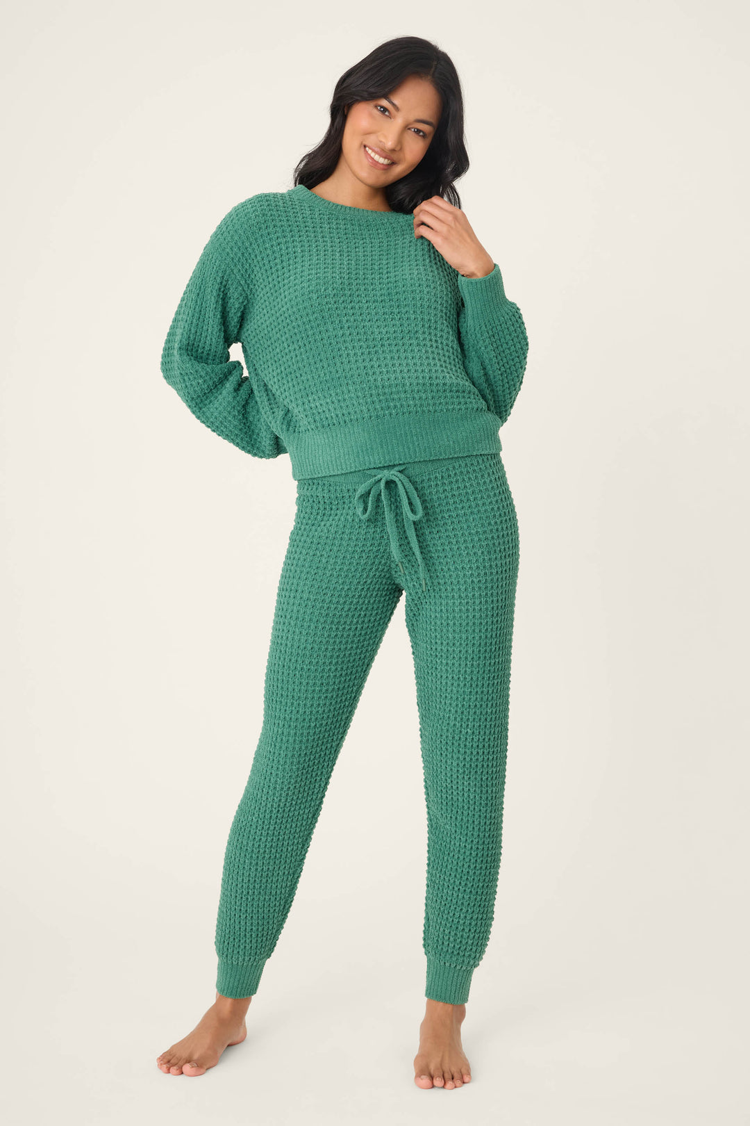 A person stands barefoot, smiling, wearing the P.J. Salvage Chenille Waffle Solid Forest Banded Pant with a matching textured green top against a plain, light background.