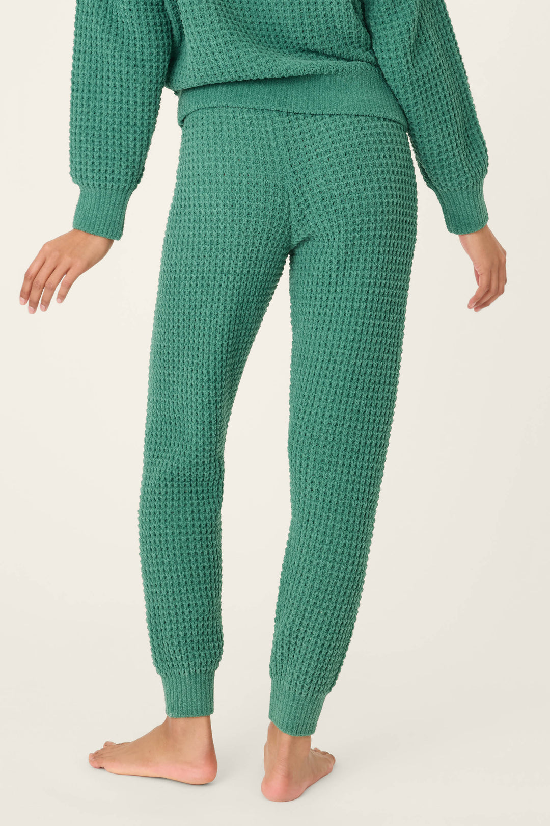 A person stands barefoot on a light surface, seen from behind wearing P.J. Salvage's Chenille Waffle Solid Forest Banded Pant and a matching sweater, highlighting the textured green knit design.