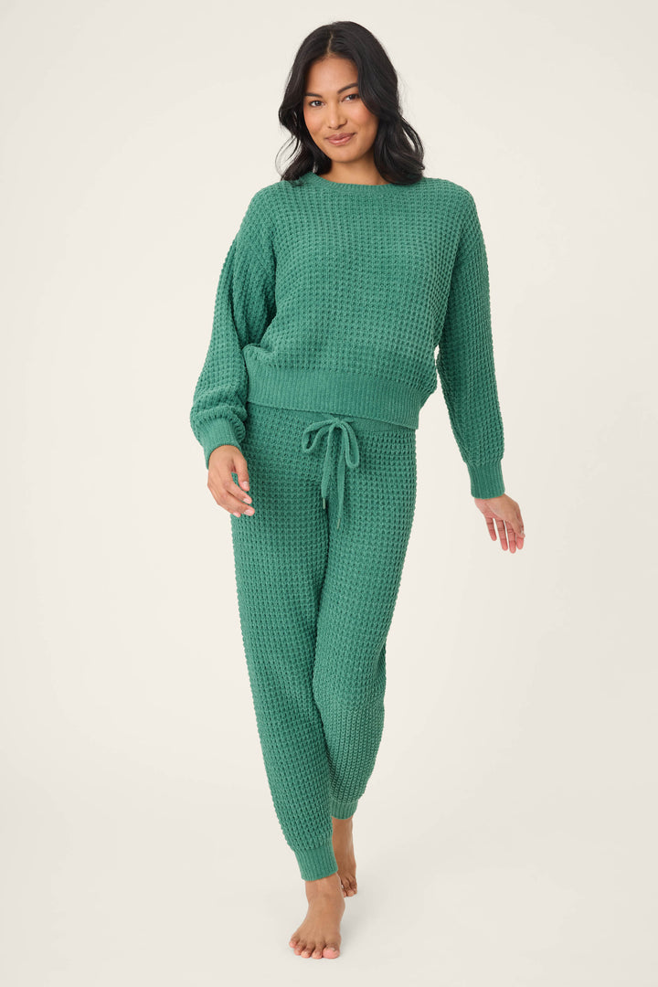 A woman with long dark hair wears the P.J. Salvage Chenille Waffle Solid Forest Lounge Set. She stands barefoot on a light background, smiling gently at the camera in the cozy green sweater and matching jogger pants.
