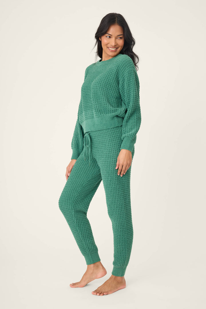 A woman smiles and looks to the side, barefoot against a plain background, wearing the P.J. Salvage Chenille Waffle Solid Forest Lounge Set with a textured green long-sleeve top and drawstring pants.