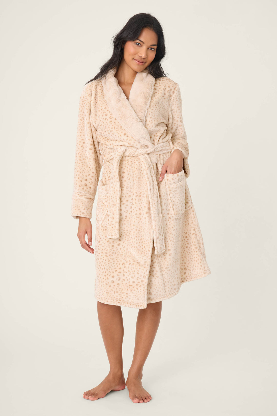 A person stands barefoot against a plain background, wearing the P.J. Salvage Luxe Plush Leopard Print Robe, a knee-length beige robe featuring an animal print, shawl collar, tie belt, and front pockets.