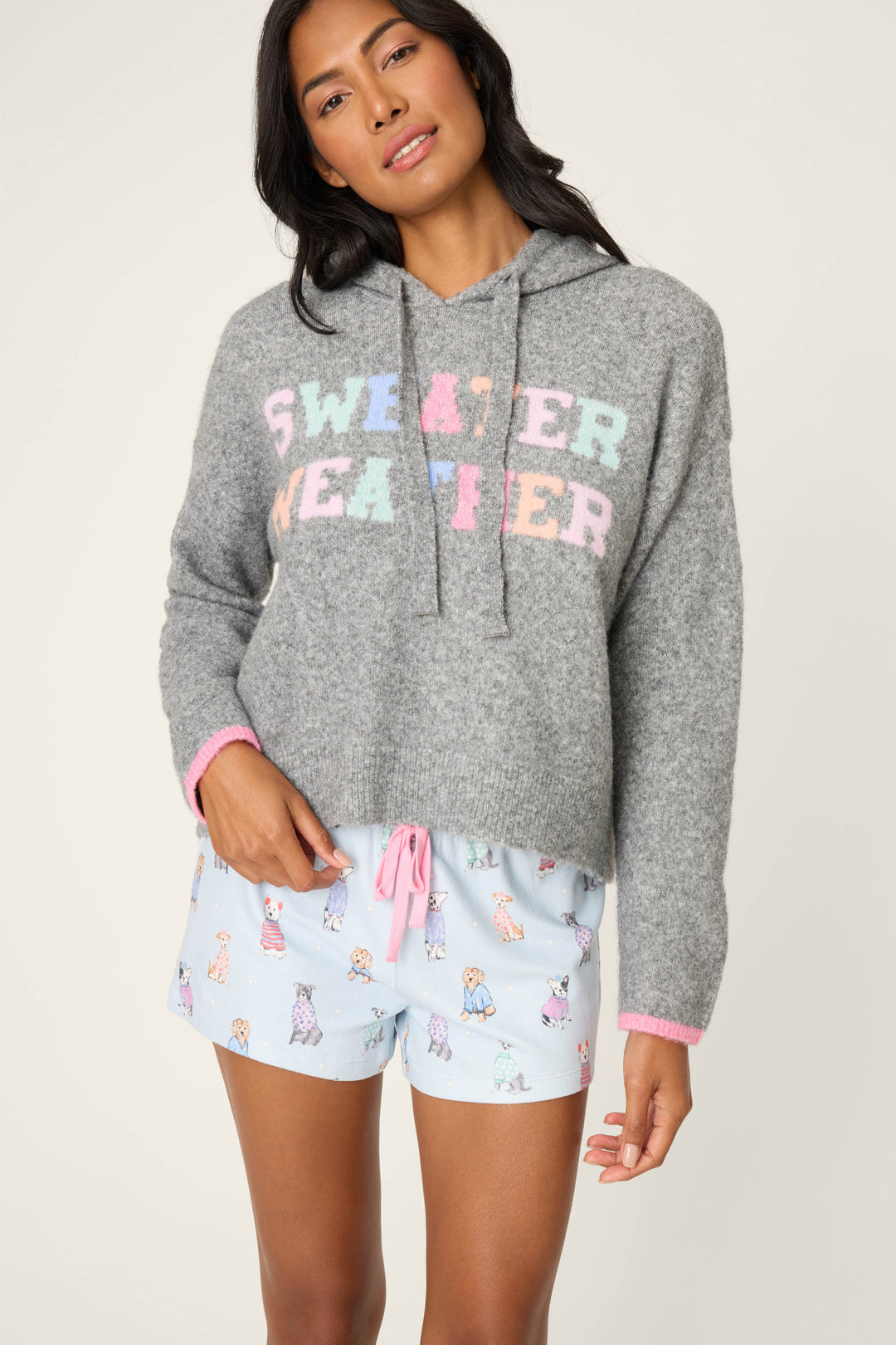 A woman poses against a plain backdrop wearing the P.J. Salvage Doggone Cold Sweater Weather Graphic Knit Hoodie and light blue pajama shorts with a cartoon dog print, with one hand in her pocket.