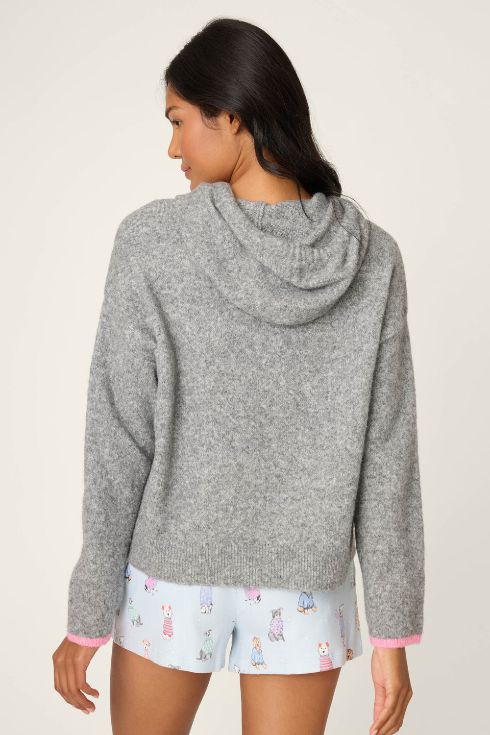 A woman with long dark hair stands with her back to the camera, wearing the P.J. Salvage Doggone Cold Sweater Weather Graphic Knit Hoodie in gray and light blue pajama shorts, against a plain light background.
