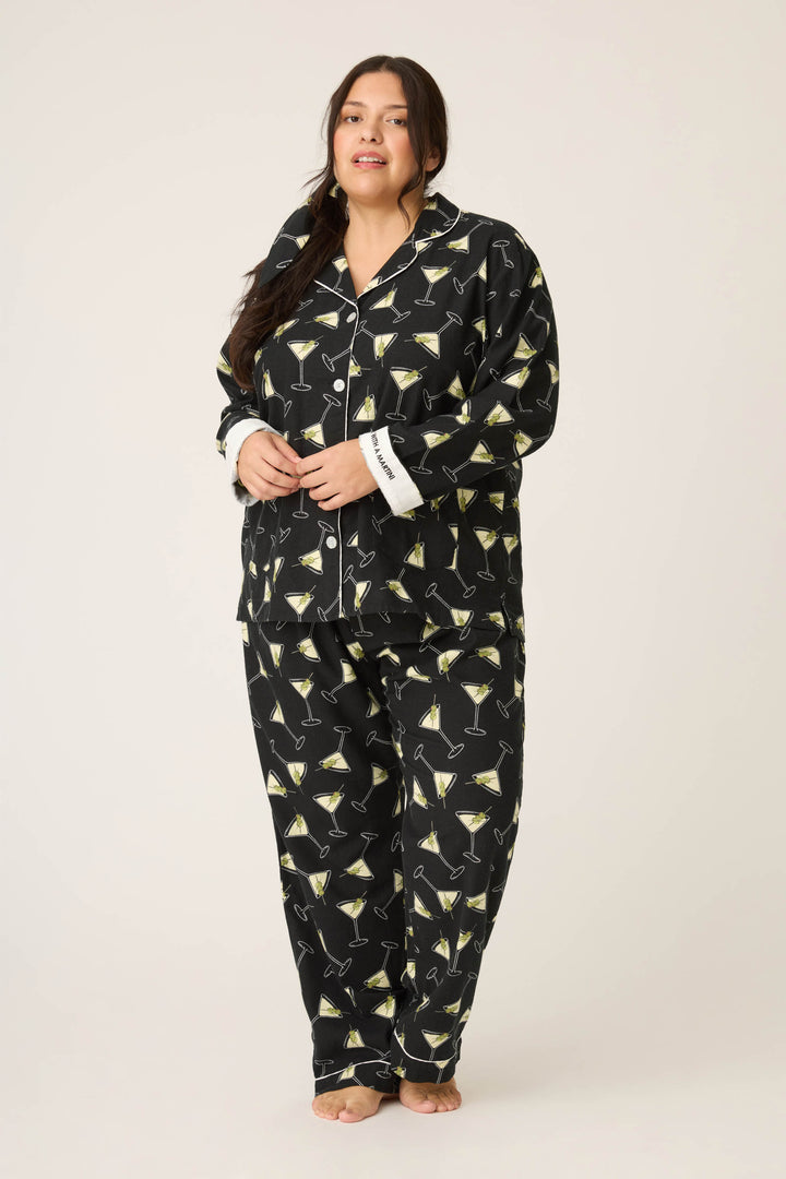A woman stands barefoot wearing the Merrier With A Martini Flannel Plus Size PJ Set by P.J. Salvage, a black pajama set featuring a martini glass print, white cuffs and buttons, with her hands loosely clasped in front of her.