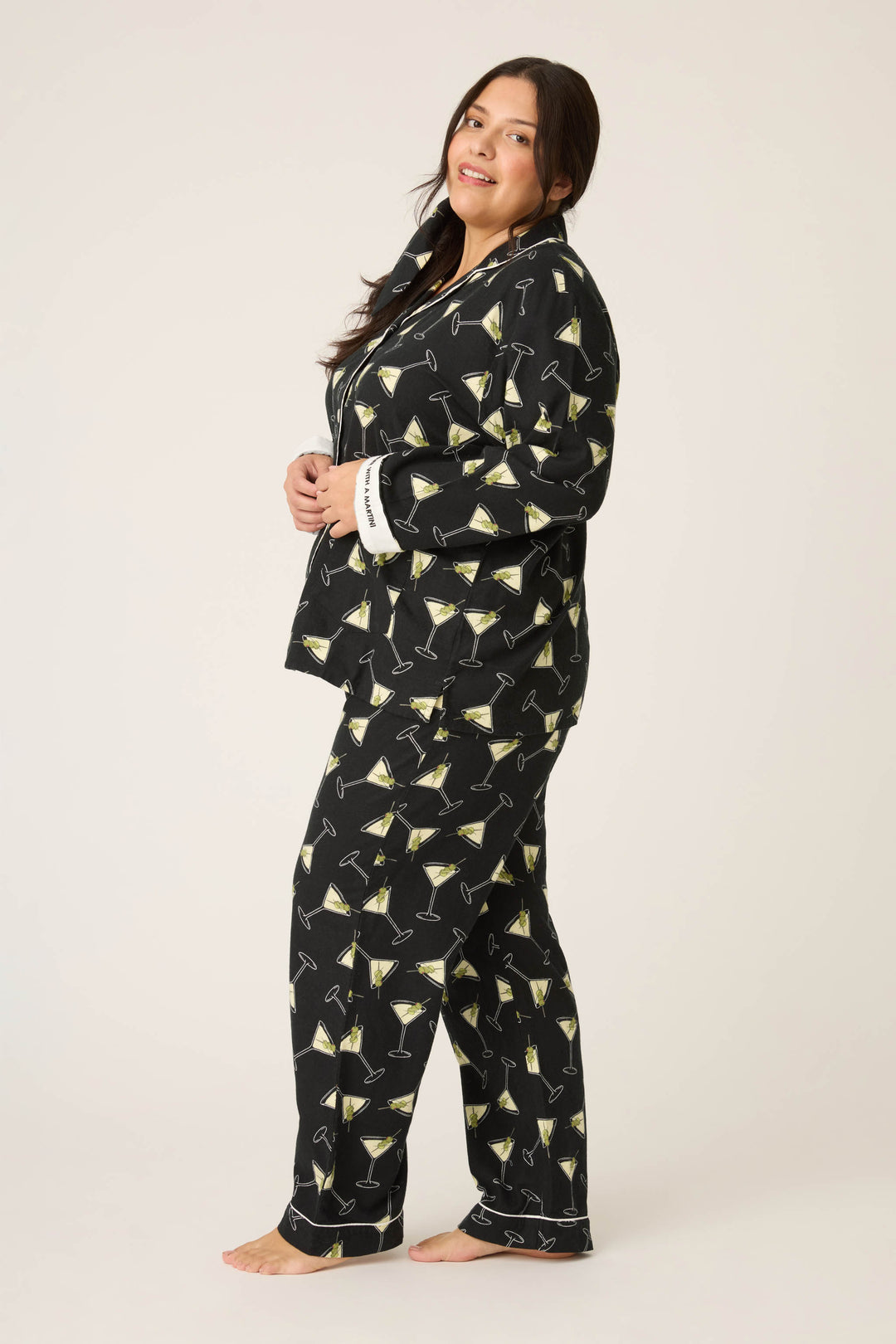 A woman stands barefoot on a plain background wearing the P.J. Salvage Merrier With A Martini Flannel Plus Size PJ Set, featuring a yellow martini glass print. She faces sideways and looks at the camera with a neutral expression.