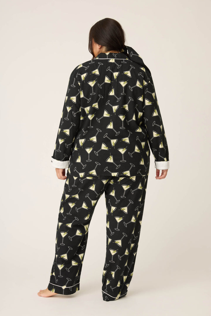 A person stands barefoot facing away, wearing the P.J. Salvage Merrier With A Martini Flannel Plus Size PJ Set featuring black pajama pants and a matching long-sleeve top, both with a repeating martini glass pattern.
