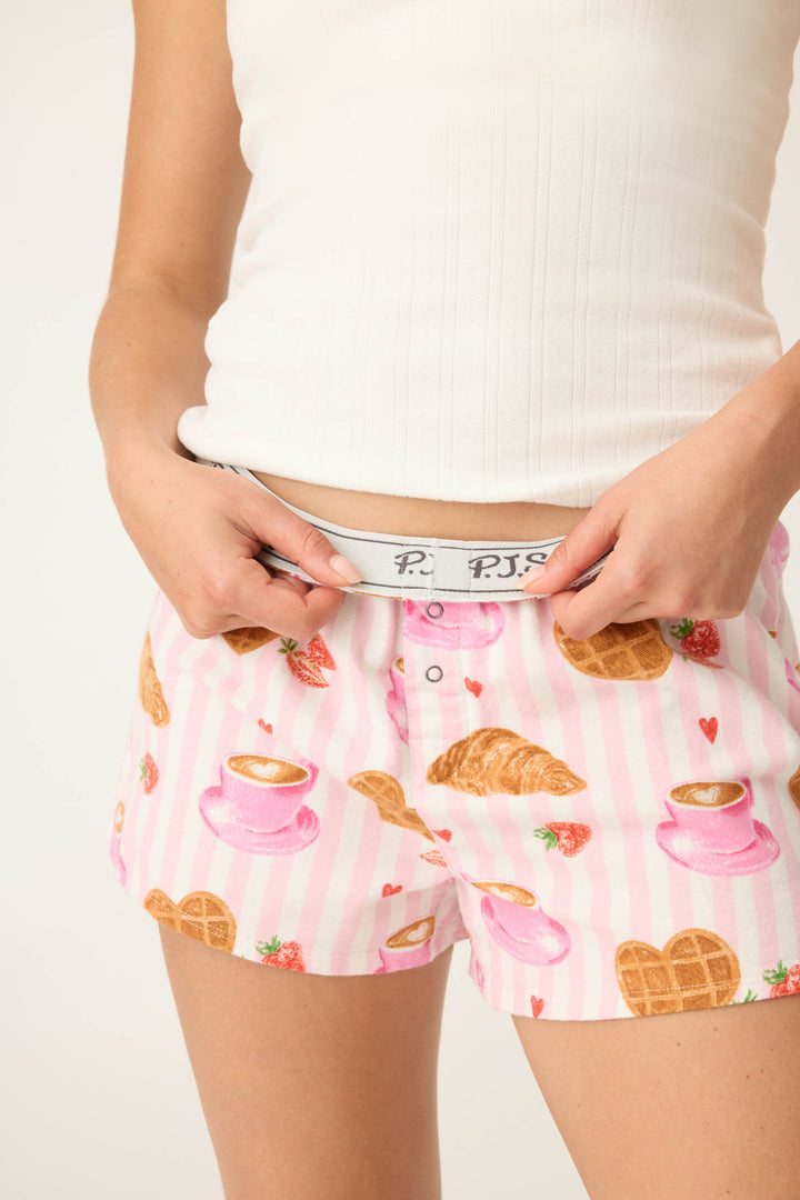 A person wearing P.J. Salvage Brunch Club Breakfast Print Flannel Shorts, featuring croissants, waffles, strawberries, and coffee cups, holds the waistband of the shorts while pairing them with a white ribbed tank top.