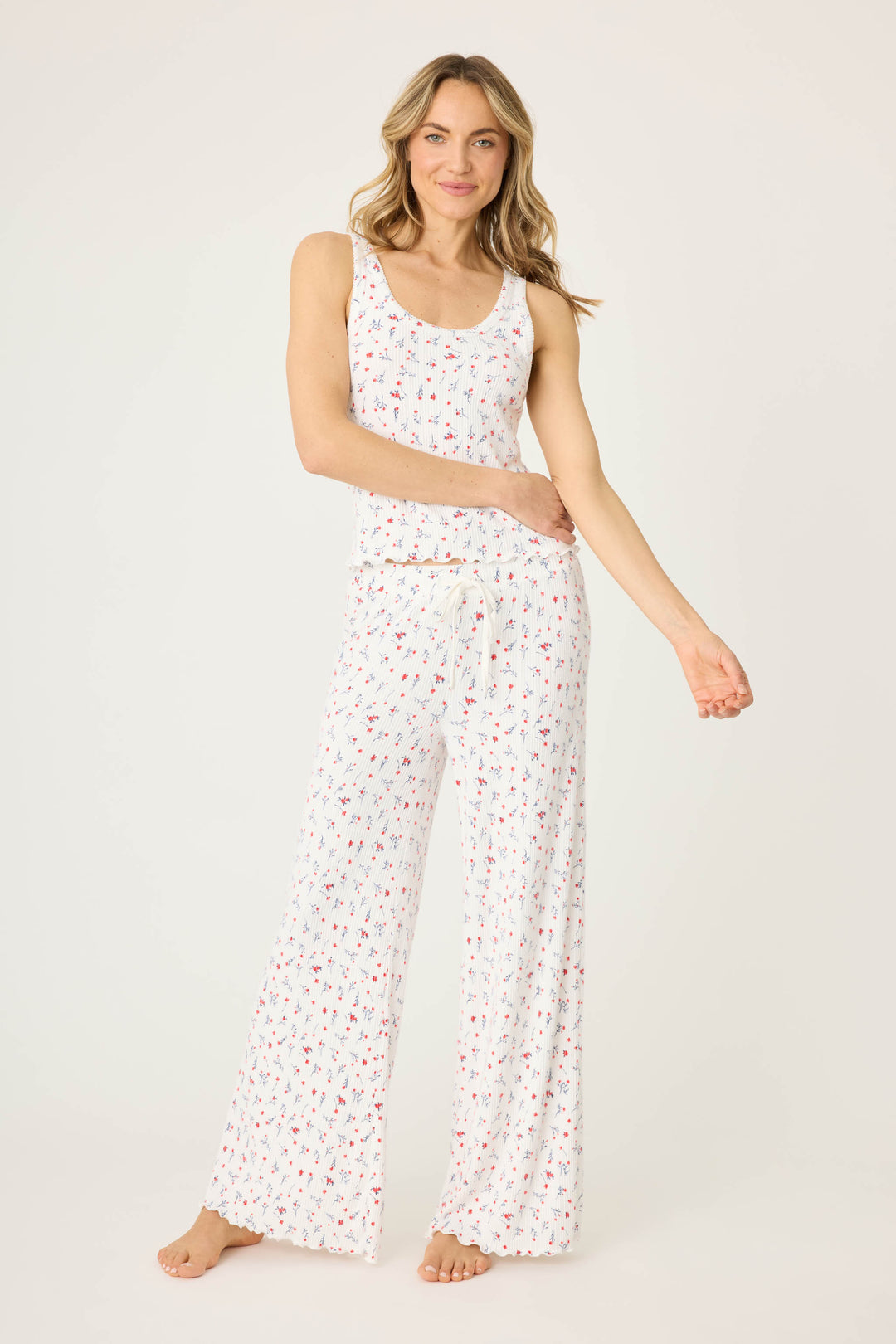 A woman with light brown hair and a relaxed expression stands barefoot on a white background, wearing the P.J. Salvage FLORAL PJ SET LA ROSE— a sleeveless white pajama set featuring small floral prints and wide-leg pants.