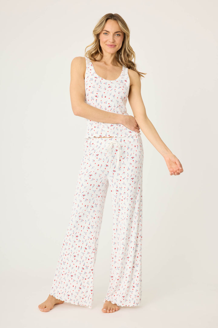 A woman with light brown hair and a relaxed expression stands barefoot on a white background, wearing the P.J. Salvage FLORAL PJ SET LA ROSE— a sleeveless white pajama set featuring small floral prints and wide-leg pants.