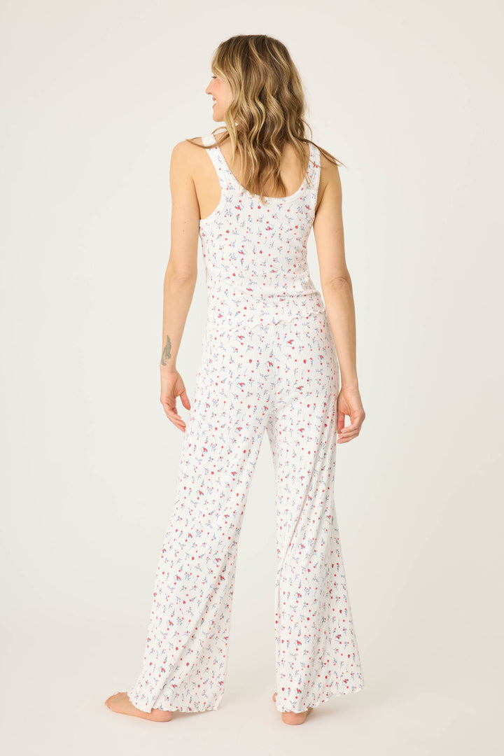 A person stands barefoot facing away, wearing the FLORAL PJ SET LA ROSE by P.J. Salvage—a sleeveless white jumpsuit with a small floral print and wide-leg pants—against a plain, light-colored background.