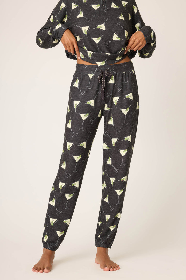 A person models the P.J. Salvage Merrier With A Martini Banded Pant—dark gray pajama pants with a martini glass print—paired with a matching top, barefoot and lifting the shirt to reveal the waistband.