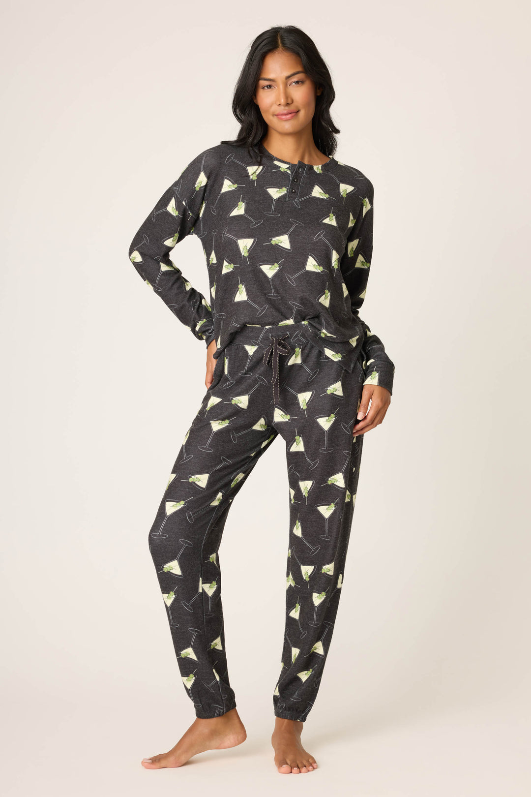 A person stands barefoot against a plain background, wearing P.J. Salvage's Merrier With A Martini Banded Pant—a matching long-sleeve pajama set with a black base and green martini glass print. One hand rests on their hip, looking relaxed.