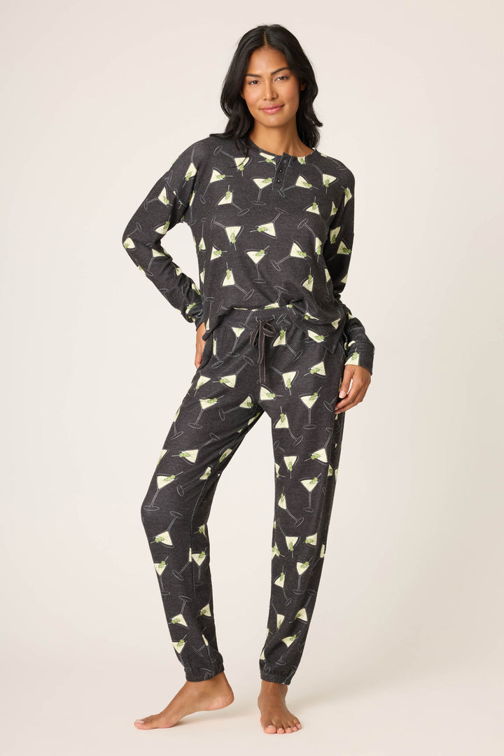 A person stands barefoot against a plain background, wearing P.J. Salvage's Merrier With A Martini Banded Pant—a matching long-sleeve pajama set with a black base and green martini glass print. One hand rests on their hip, looking relaxed.