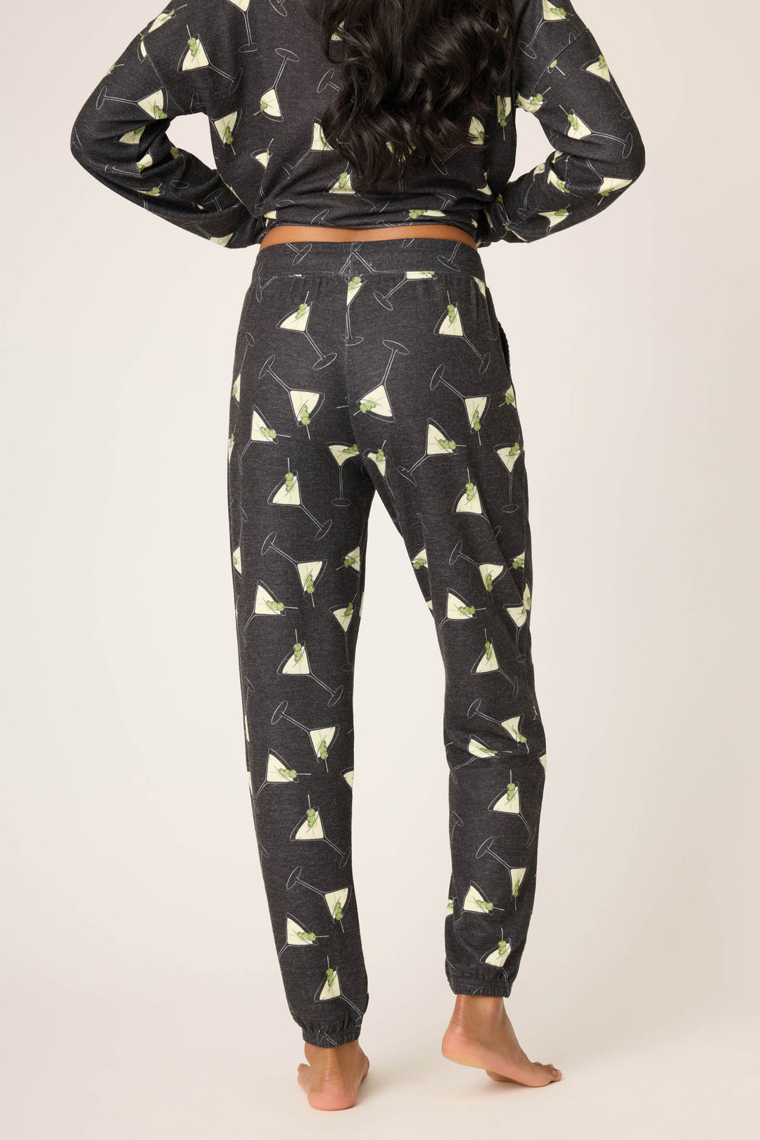 A person stands barefoot, facing away, wearing P.J. Salvage Merrier With A Martini Banded Pant and matching top with a martini glass pattern. The background is plain and light-colored.