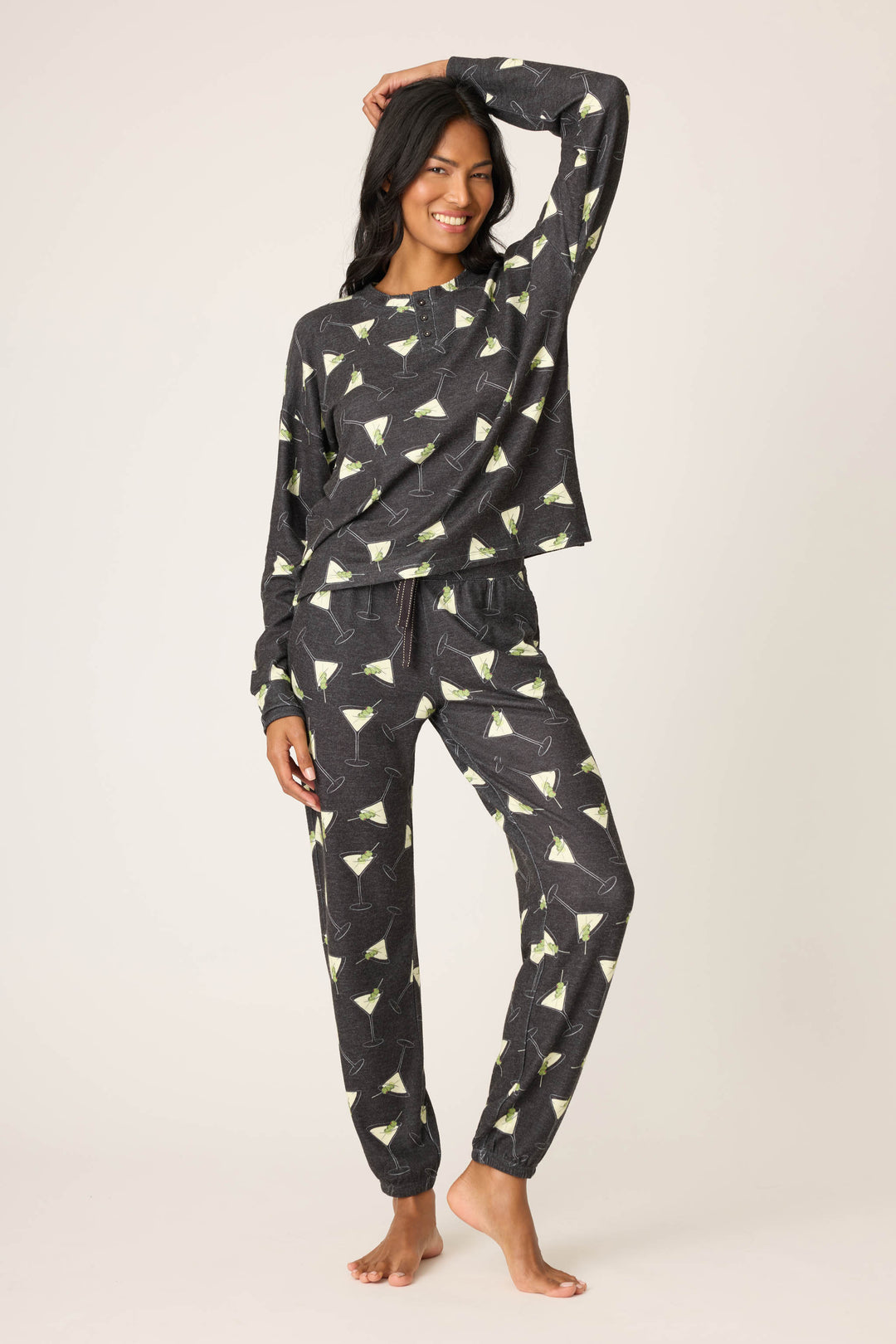 A woman smiles barefoot with one hand on her head, wearing the P.J. Salvage Merrier With A Martini PJ Set—a dark, long-sleeve top and pants featuring a martini glass print—against a plain, light background.
