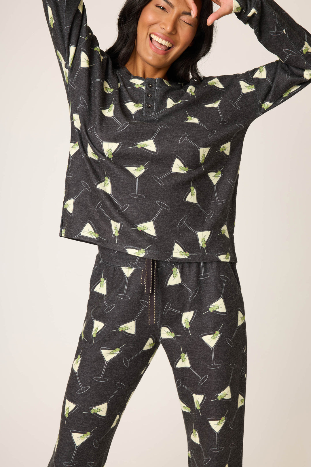 A person smiles and poses in the P.J. Salvage Merrier With A Martini PJ Set, featuring dark pajama pants and a long-sleeve top with a repeating martini glass print, raising one hand near their face.