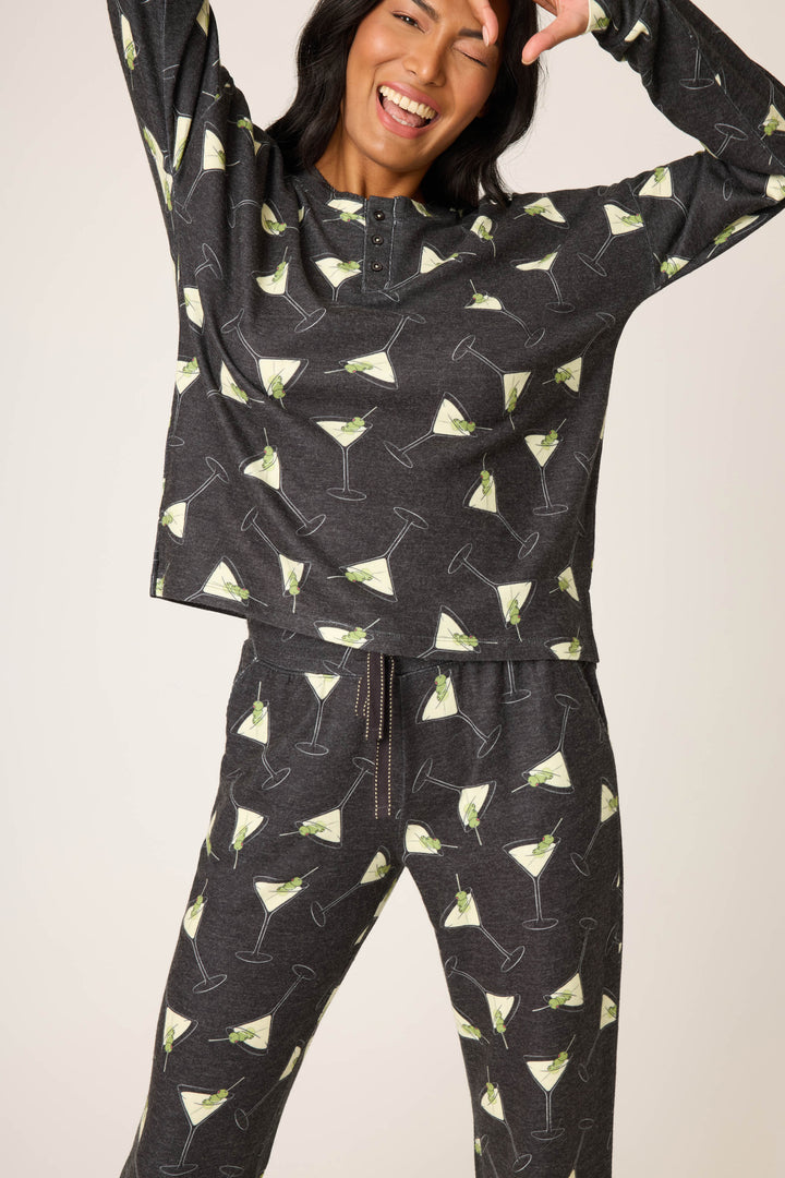 A person smiles and poses in the P.J. Salvage Merrier With A Martini PJ Set, featuring dark pajama pants and a long-sleeve top with a repeating martini glass print, raising one hand near their face.