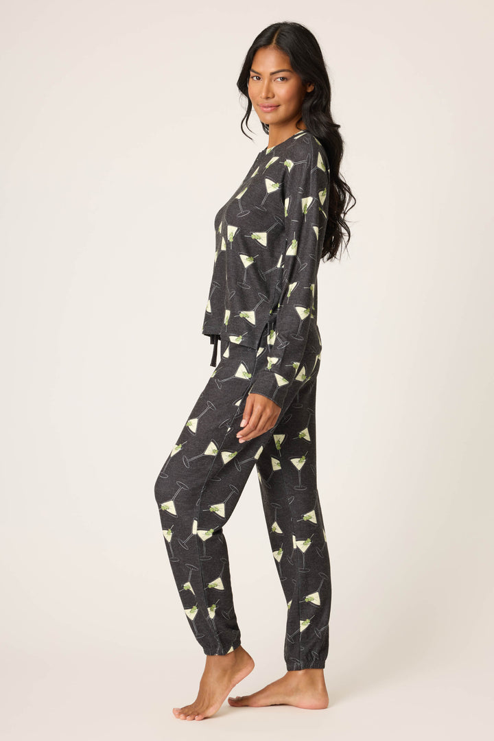 A woman, barefoot and smiling softly, wears the P.J. Salvage Merrier With A Martini PJ Set—a matching dark long-sleeve top and jogger pants with a martini glass print—while standing sideways.