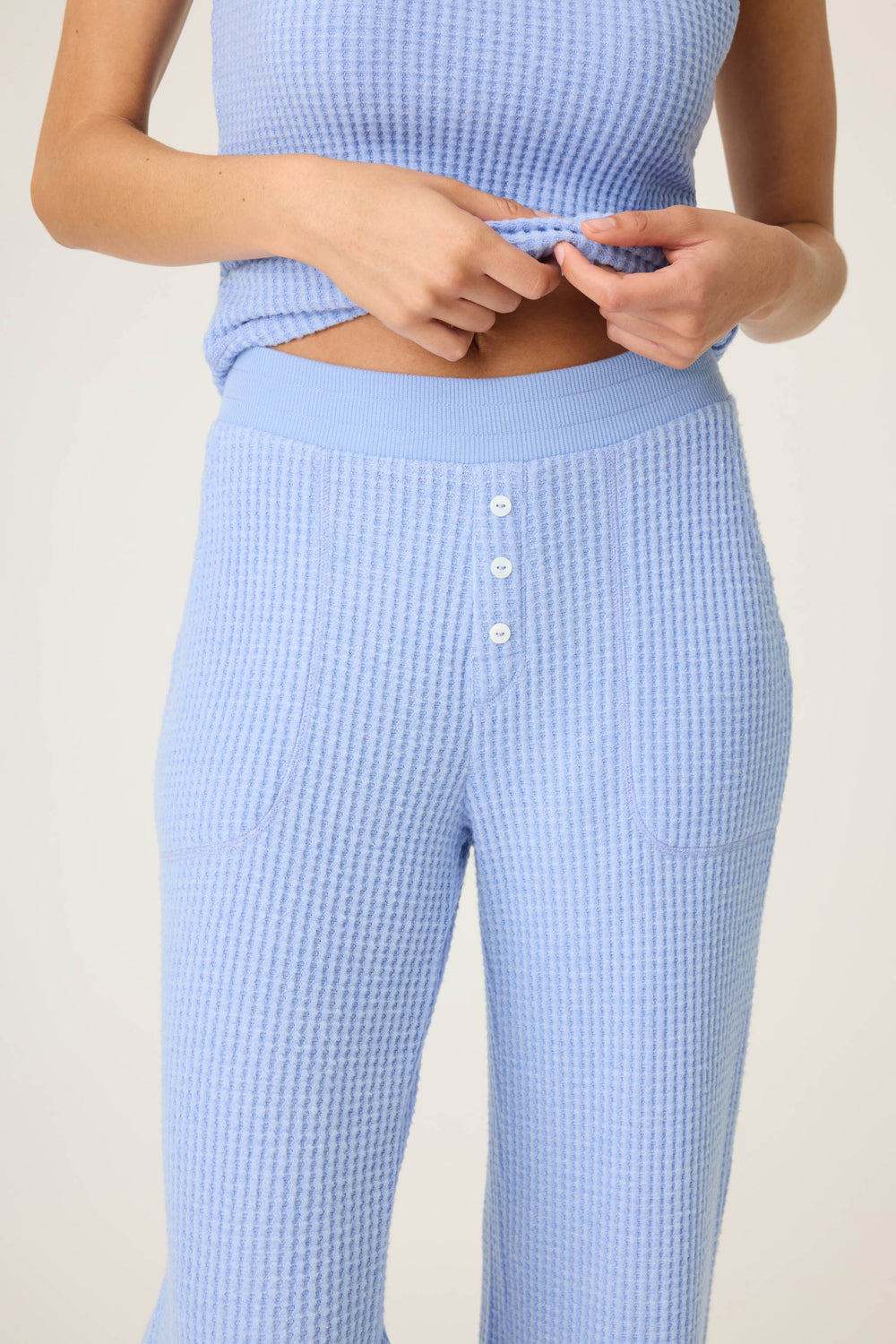 A person is shown from torso to upper legs wearing the P.J. Salvage Morning Waffles Solid Peri Open Leg Pant and a matching light blue, sleeveless, ribbed top with three white buttons, holding the hem of the top in both hands.
