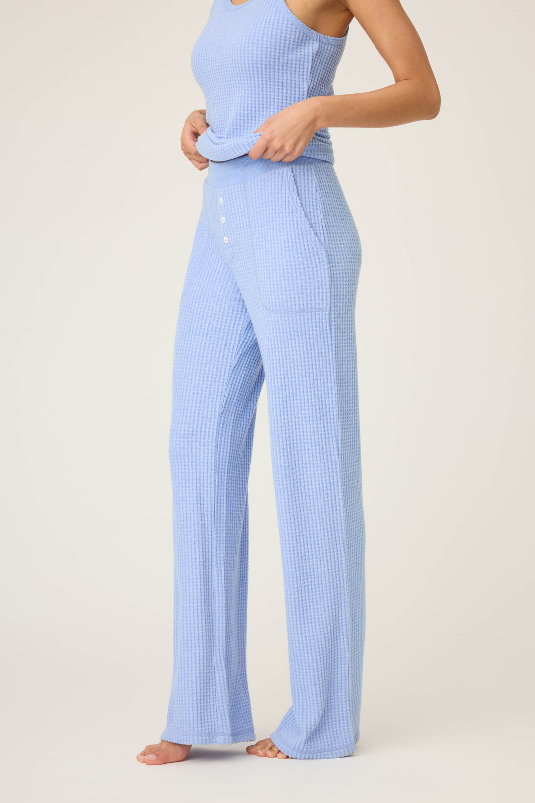A person stands barefoot against a light background, adjusting their top while wearing P.J. Salvage’s Morning Waffles Solid Peri Open Leg Pant—light blue, high-waisted, wide-leg pants featuring front button details.
