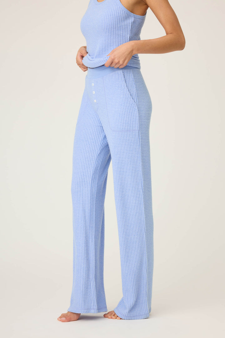 A person stands barefoot against a light background, adjusting their top while wearing P.J. Salvage’s Morning Waffles Solid Peri Open Leg Pant—light blue, high-waisted, wide-leg pants featuring front button details.