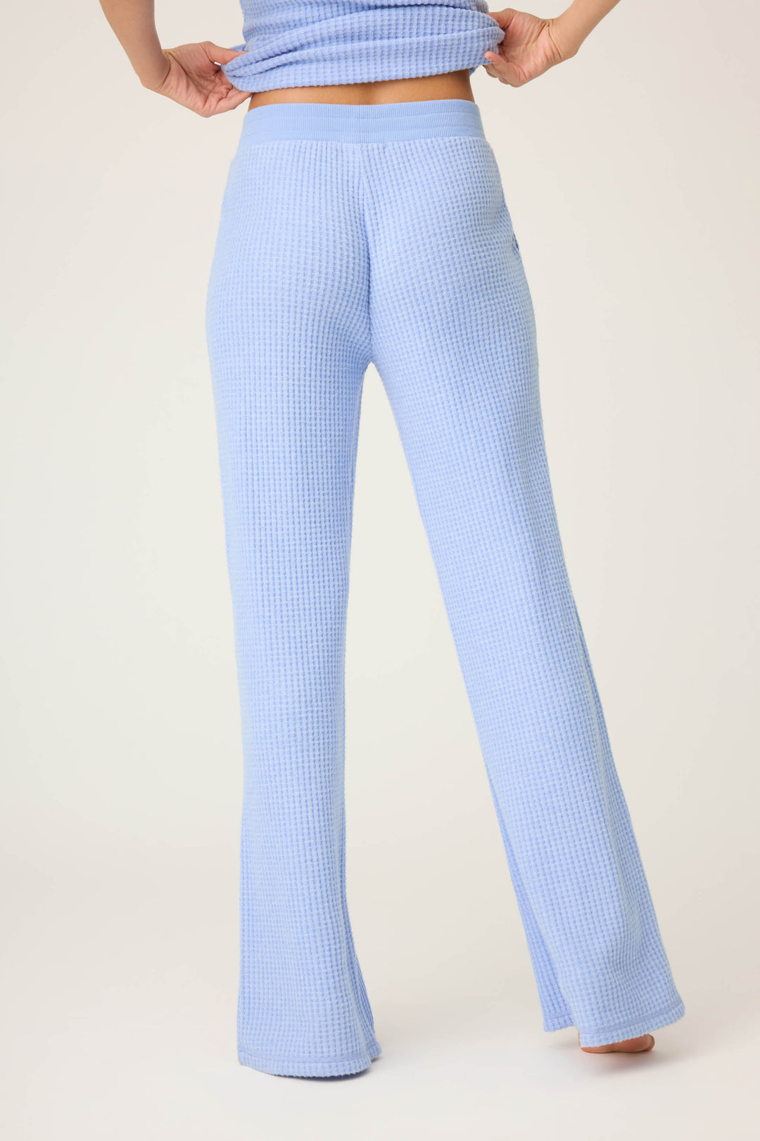 A person stands with hands on hips, wearing P.J. Salvage’s Morning Waffles Solid Peri Open Leg Pant, featuring a high-waisted, textured light blue design with a slight flare, photographed from the back against a plain background.