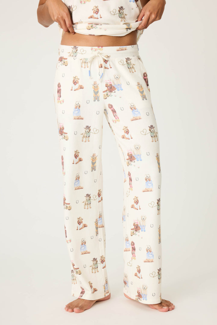 A person stands barefoot in P.J. Salvage’s DOGS OPEN LEG PANT RUFF ON THE RANGE, white pajama pants with a drawstring waist and playful pattern, holding up the matching top that is partially visible.