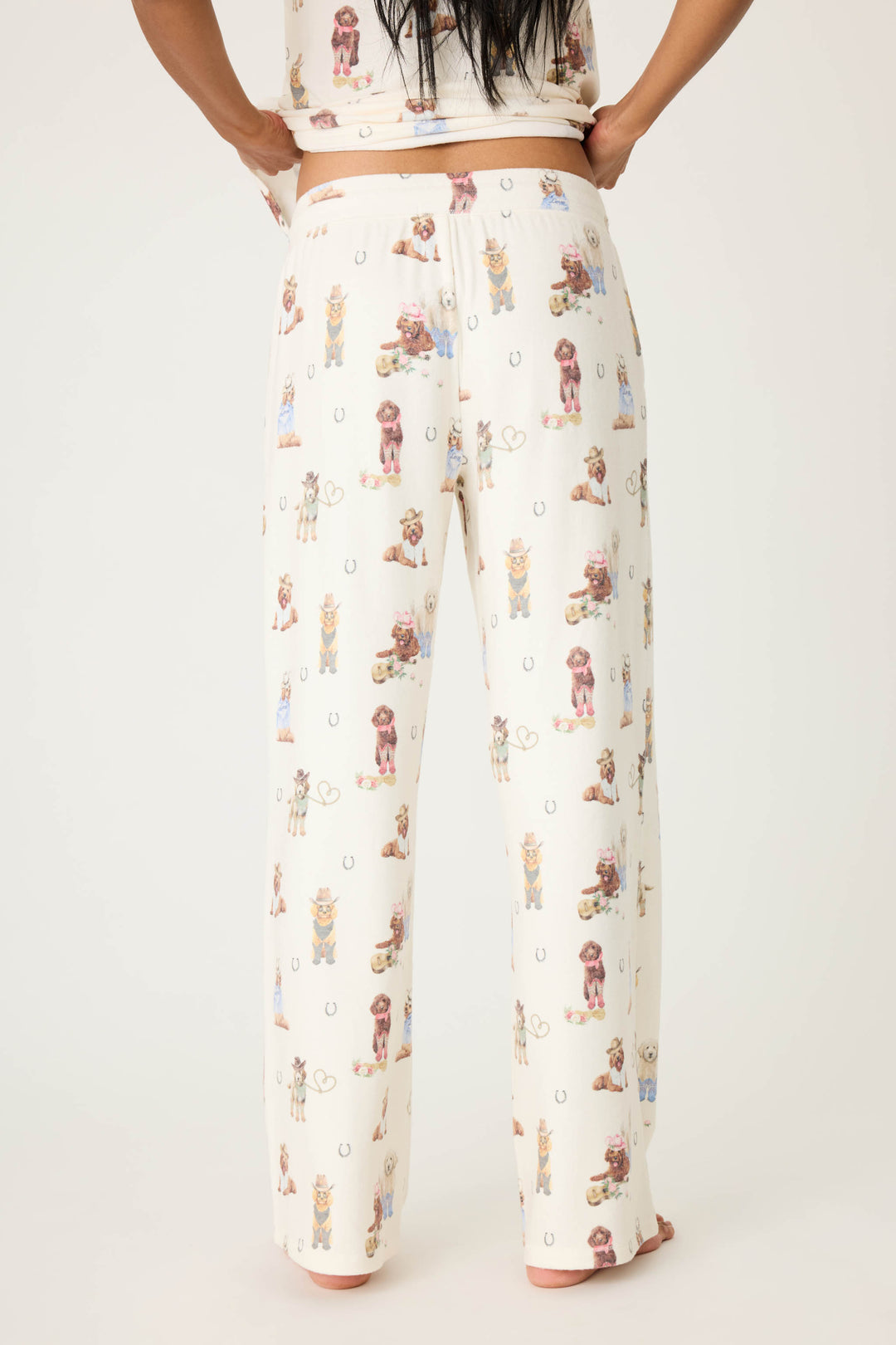 A person is shown from the back, barefoot against a plain light background, wearing P.J. Salvage DOGS OPEN LEG PANT RUFF ON THE RANGE—a cream-colored pajama pant with an all-over dogs and horseshoes print.