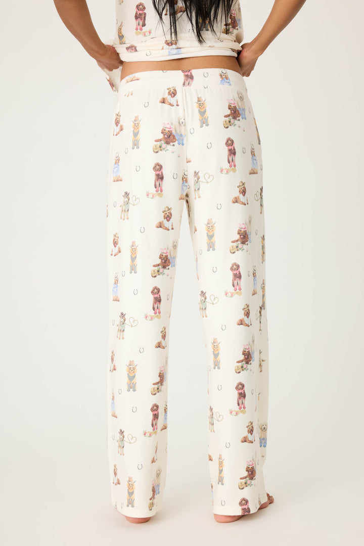 A person is shown from the back, barefoot against a plain light background, wearing P.J. Salvage DOGS OPEN LEG PANT RUFF ON THE RANGE—a cream-colored pajama pant with an all-over dogs and horseshoes print.