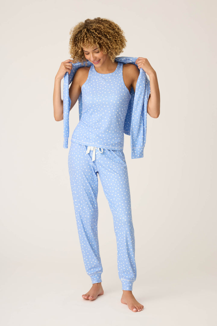 A person stands barefoot in P.J. Salvage’s Pointelle Essentials Ditsy Star 4-Piece Loungewear Set, featuring a light blue tank top and drawstring pants with white stars, holding the matching top against a plain, light background.