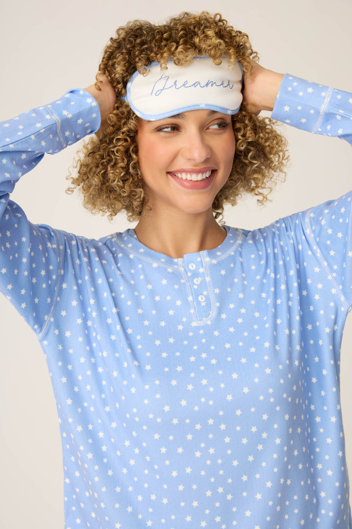 A person in the P.J. Salvage Pointelle Essentials Ditsy Star 4-Piece Loungewear Set smiles while holding a blue-and-white “Dreamer” sleep mask on their forehead.