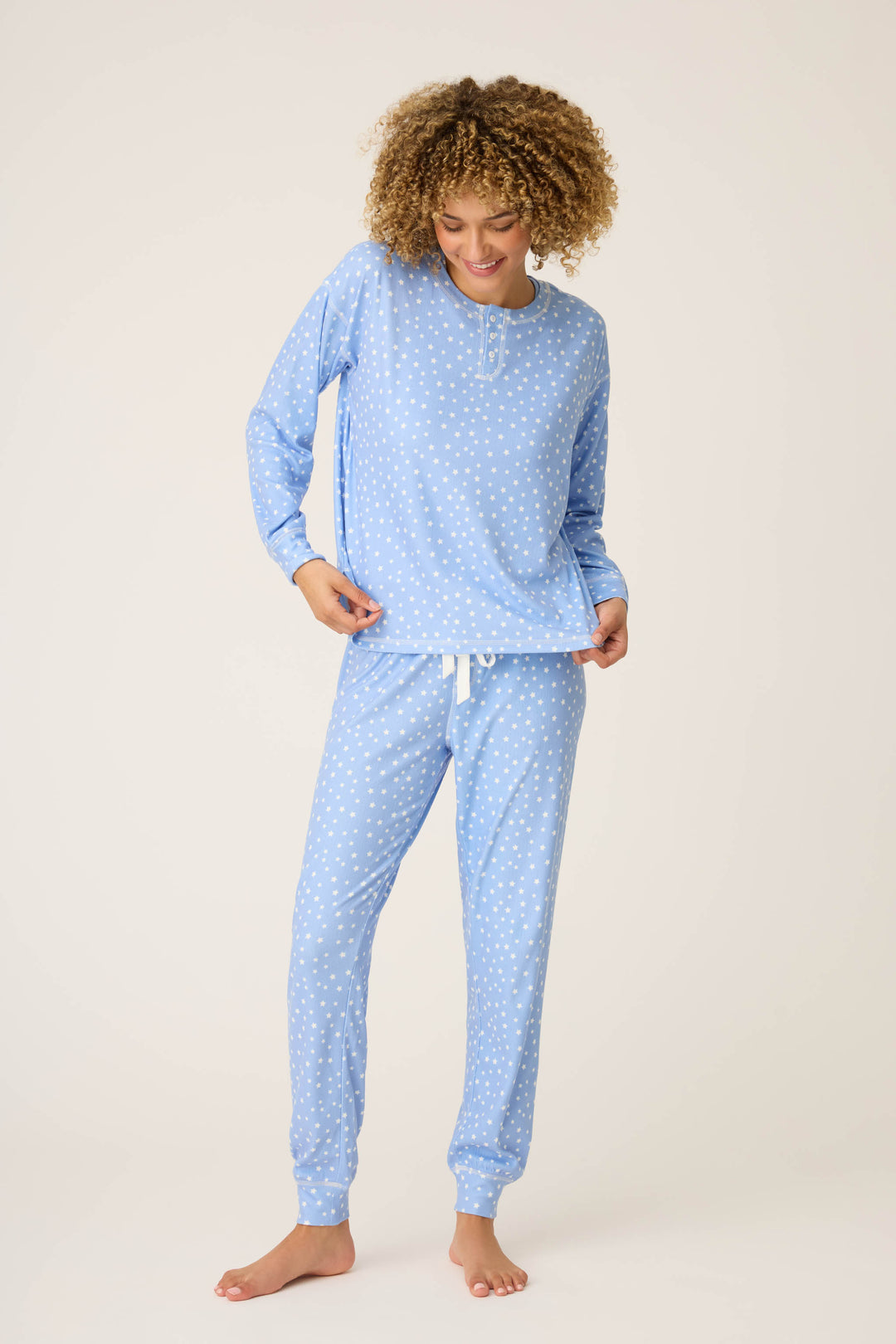 A person stands barefoot against a plain background, wearing the P.J. Salvage Pointelle Essentials Ditsy Star 4-Piece Loungewear Set—light blue, with a long-sleeve top and drawstring pants. They look down and smile slightly.