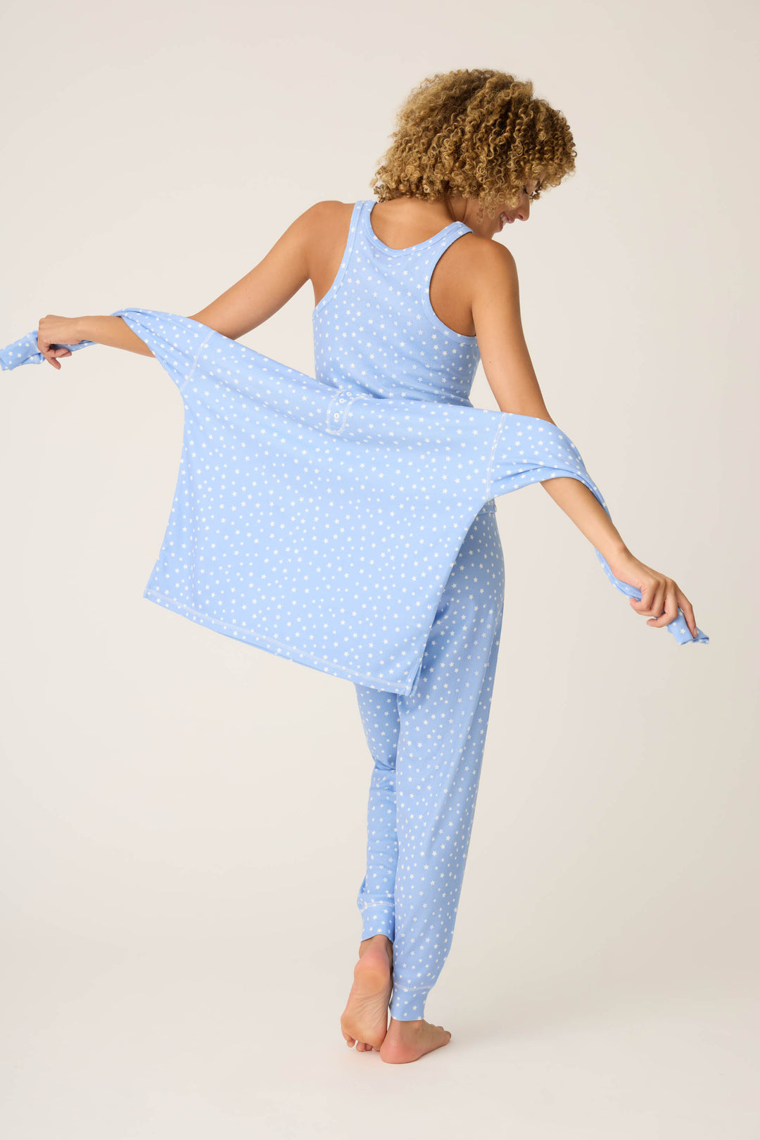 A person stands barefoot, facing away, wearing the P.J. Salvage Pointelle Essentials Ditsy Star 4-Piece Loungewear Set in light blue and holding a matching robe spread out behind their back.