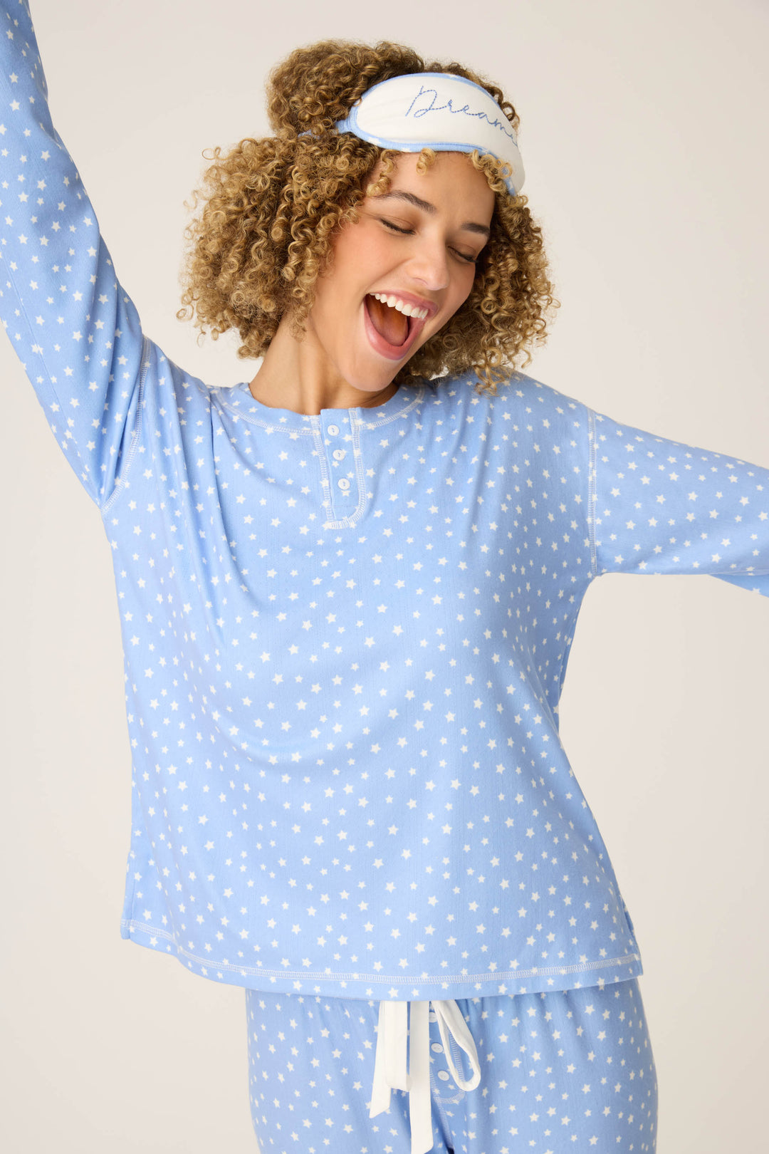 A person wearing the P.J. Salvage Pointelle Essentials Ditsy Star 4-Piece Loungewear Set smiles joyfully with arms raised and a white sleep mask, standing against a plain background.
