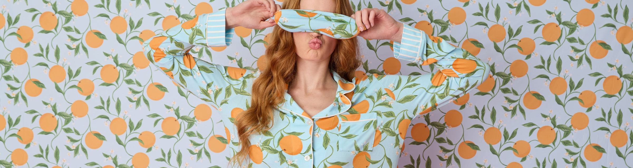 A woman wearing maroon pajamas stands next to a wooden rod displaying five pairs of colorful pajama pants with various patterns, against a beige background.