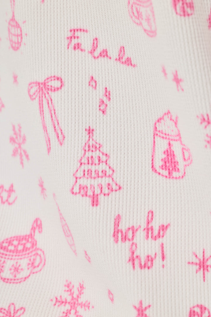 A close-up of a white knit fabric with a pink holiday print featuring a Christmas tree, hot cocoa cups, snowflakes, bows, ornaments, and festive text.